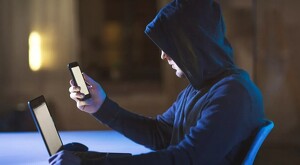 Hombre al que no se le ve el rostro vestido con una sudadera con capucha volteando a ver un celular y con una laptop enfrente