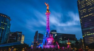 Vista de noche del monumento del Ángel de la Independencia en Ciudad de México con luces azules y rosas iluminándolo y edificios alrededor