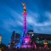 Vista de noche del monumento del Ángel de la Independencia en Ciudad de México con luces azules y rosas iluminándolo y edificios alrededor