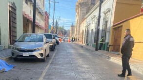 Se observa a un hombre portando un uniforme de policía y a una persona tirada en el suelo con una sábana azul encima; se le ven unso tenis.