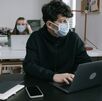 Un hombre joven en una oficina en compañía de una mujer joven que usan un cubrebocas mientras están trabajando frente a su computadora portátil