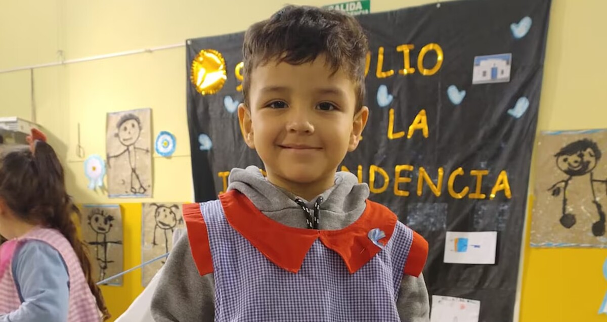 Niño Valentín Mercado Toledo sonriendo mientras ve a la cámara con un mandil de cuadros dentro de un salón de kínder con una niña a un lado y dibujos de fondo