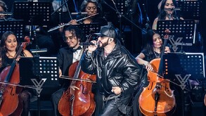 Se observa a un hombre con chamarra negra y a unas personas al fondo tocando instrumentos musicales.