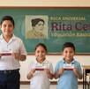 Pizarrón con la imagen de la beca Rita cetina y  en frente hay tres niños en un salón de clases que sostienen una tarjeta del bienestar