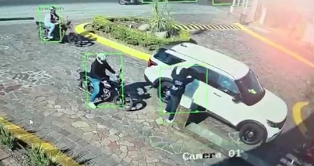 Dos motociclistas y un hombre golpeando el vidrio de una camioneta blanca a las afueras de un fraccionamiento con plantas y empedrado