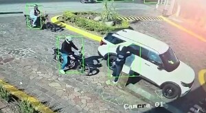 Dos motociclistas y un hombre golpeando el vidrio de una camioneta blanca a las afueras de un fraccionamiento con plantas y empedrado