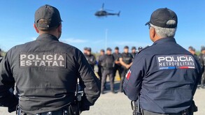 Oficial de la Policía de Jalisco al lado de un elemento de la Policía Metropolitana parados delante de policías del estado con un helicóptero sobrevolando
