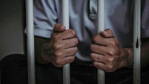 Hombre visto del toros hacia abajo adentro de una prisión sosteniendo los barrotes de una celda y vistiendo una camisa tipo polo color blanca