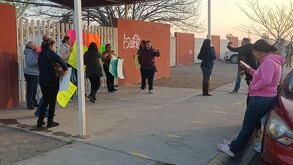Se observa a varias personas paradas, afuera de una escuela, con pancartas en mano.