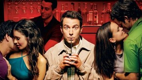 Hombre viendo a la cámara con expresión de sorpresa mientras bebe de un popote estando en medio de dos parejas besándose en un bar