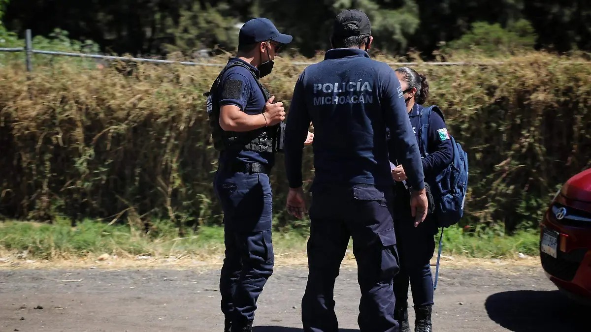 Dos hombres y una mujer miembros de la Policía de Michoacán parados en medio de una calle con pastizales al fondo y un auto rojo a un costado