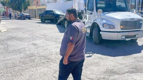 Hay una camioneta negra al fondo, una grúa y un hombre que viste playera gris y pantalón negro.