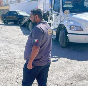 Hay una camioneta negra al fondo, una grúa y un hombre que viste playera gris y pantalón negro.