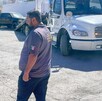 Hay una camioneta negra al fondo, una grúa y un hombre que viste playera gris y pantalón negro.