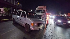 Camioneta tipo van color gris de la marca Ford con parte del fascia destruida por impacto con un tráiler que está detrás con autos transitando alrededor