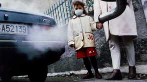 Una niña usando cubrebocas acompañada por su madre frente a un auto que libera contaminantes del escape