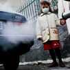 Una niña usando cubrebocas acompañada por su madre frente a un auto que libera contaminantes del escape