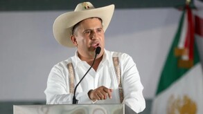 Carlos Alberto Manzo Rodríguez hablando por un micrófono ante un podio con una bandera de México difuminada en el fondo durante presentación pública