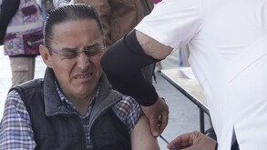 Se observa a un hombre haciendo gestos y viste de camisa de cuadros con chaleco gris con negro.