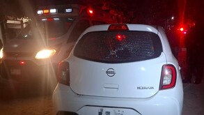 Vista de la parte trasera de un carro Nissan March color blanco con el cristal roto por impactos de bala y una ambulancia estacionada a un lado