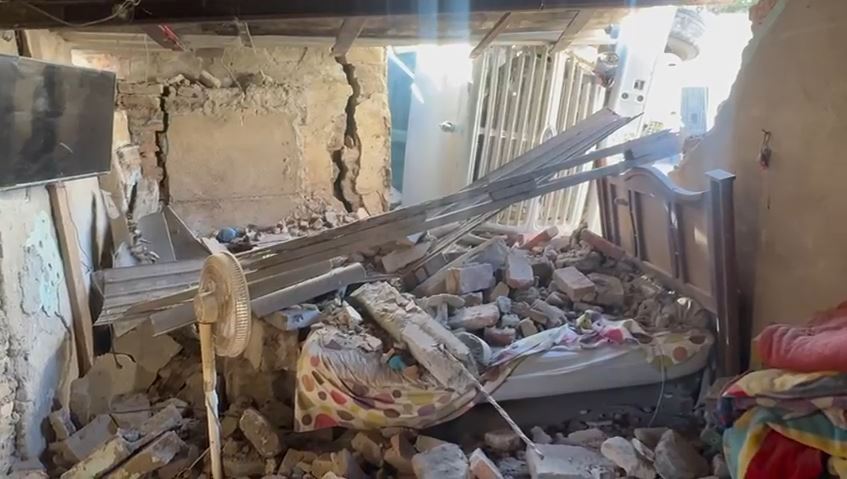 Láminas, escombros y ladrillos esparcidos sobre una cama con un ventilador y una tele al interior de una casa con las paredes destruidas