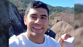 Fernando Serrano sonriendo mientras mira de frente a la cámara con un paisaje con piedras y árboles de fondo con una mujer a un lado