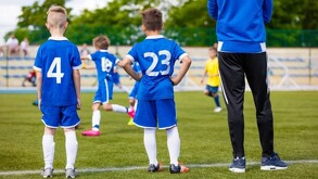 Hombre parado al lado de dos niños observando un partido de fútbol infantil en una cancha de pasto con gradas y familias desenfocadas en el fondo