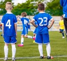 Hombre parado al lado de dos niños observando un partido de fútbol infantil en una cancha de pasto con gradas y familias desenfocadas en el fondo