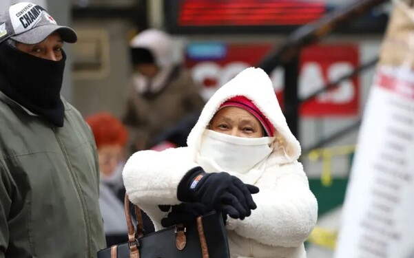 Adultos mayores cubiertos de los ojos hacia abajo y vistiendo abrigos, gorra y guantes con personas desenfocadas en el fondo