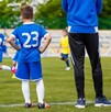 Hombre parado al lado de dos niños observando un partido de fútbol infantil en una cancha de pasto con gradas y familias desenfocadas en el fondo