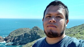 Selfie de Alejandro Garín viendo a la cámara delante del paisaje de un acantilado frente al mar con el cielo despejado
