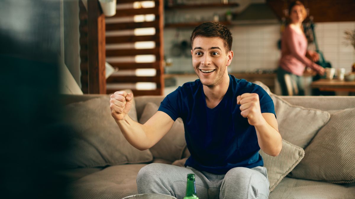 Hombre viendo el futbol en la sala mientras su esposa lo observa desde atrás.