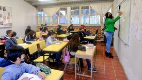 Maestra escribiendo en una pizarra blanca delante de un grupo de alumnos de educación básica adentro de un salón de clases