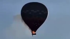 Globo aerostático visto a contraluz en pleno vuelo con fuego en la canastilla, humo alrededor y la silueta de una persona cayendo