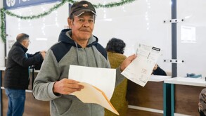 Hombre mayor sosteniendo carpetas y documentos con una media sonrisa en el rostro delante de ventanillas en una oficina recaudadora