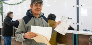 Hombre mayor sosteniendo carpetas y documentos con una media sonrisa en el rostro delante de ventanillas en una oficina recaudadora