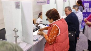 Mujer mayor con documentos en la ventanilla de una oficina recaudadora en Zapopan con personas en el fondo desenfocadas