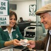 Adulto mayor sonriendo mientras recibe el pago de la pensión del IMSS a través de una ventanilla con una mujer haciendo la entrega de los billetes