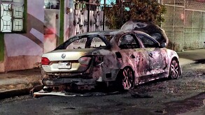 Automóvil Volkswagen Jetta color gris calcinado y con el cofre levantado estacionado al exterior de una casa y con rastros de incendio en la calle