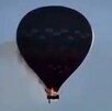 Globo aerostático visto a contraluz en pleno vuelo con fuego en la canastilla, humo alrededor y la silueta de una persona cayendo
