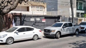 Camioneta gris y autos blancos estacionados al exterior de una vivienda con portón negro y sellos de clausura puestos