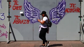 Mujer caminando mientras ve su celular con una valla pintada con mensajes y unas alas moradas detrás