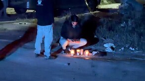 Mujer en cuclillas viendo su teléfono delante de unas veladoras colocadas sobre una calle con un hombre parado al lado de ella