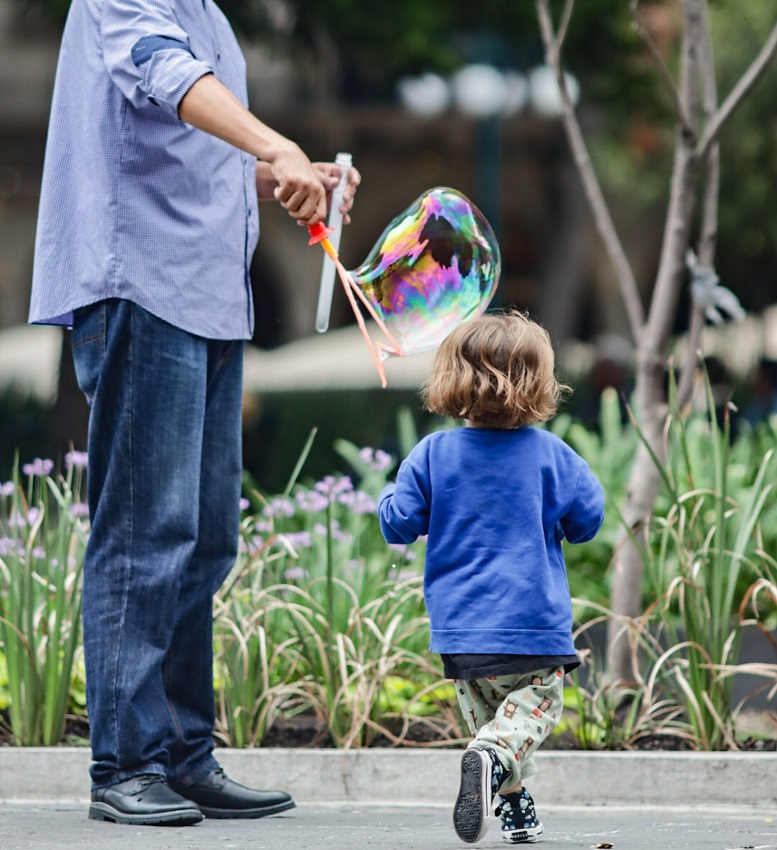Hombre alto juega a lar burbujas con un niño rubio en una plaza.