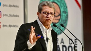 Hombre realiza gesto con la boca y alza la mano mientras habla ante un micrófono.