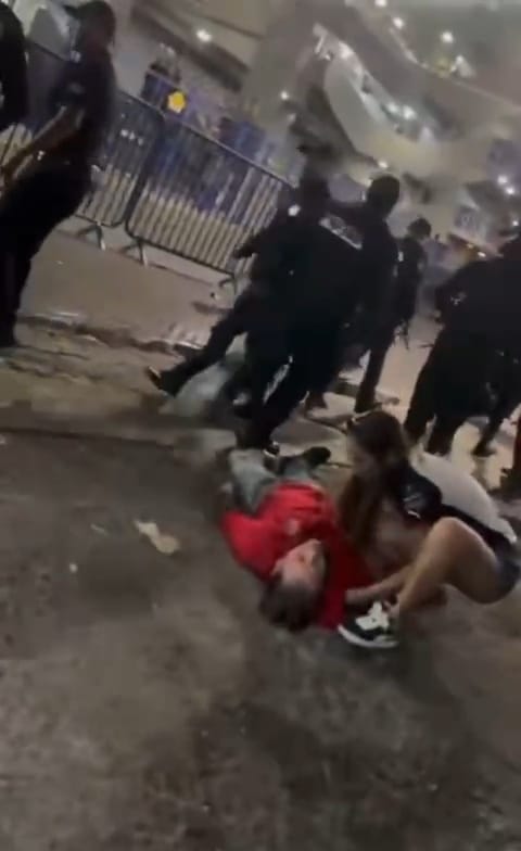 Hombre aficionado de Atlas tendido inconsciente sobre el suelo con una mujer tratando de levantarlo y policías alrededor