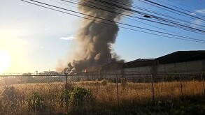 Columna de humo gris emanando del interior de una fábrica incendiándose vista desde lejos con una malla metálica de seguridad alrededor