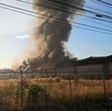 Columna de humo gris emanando del interior de una fábrica incendiándose vista desde lejos con una malla metálica de seguridad alrededor