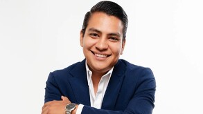 Se observa a un hombre sonriendo, con traje color azul marino y está con los brazos cruzados; trae un reloj.