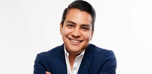 Se observa a un hombre sonriendo, con traje color azul marino y está con los brazos cruzados; trae un reloj.